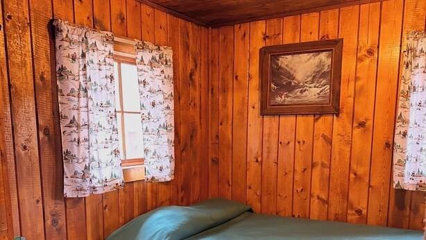 A small, cozy room with wooden walls, a single bed, a chair, and a framed picture on the wall.