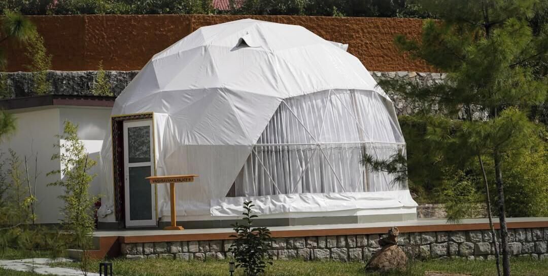 Una estructura tipo tienda blanca con una puerta y una pequeña mesa en frente, ubicada contra un muro de piedra y rodeada de follaje.