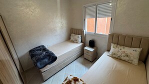 A bedroom with a single bed, a smaller bed, a bedside table, a window with blinds, and a ceiling light. - Charming 3-Bedroom Apartment Near Menara Garden (Marrakesh)