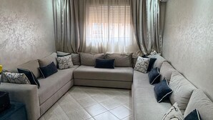 A modern living room with a sectional sofa, a chandelier, and a large window with curtains. - Charming 3-Bedroom Apartment Near Menara Garden (Marrakesh)