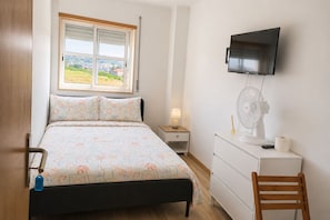Ein Schlafzimmer mit einem Bett, einem Nachttisch mit Lampe, einem an der Wand montierten Fernseher, einem Ventilator und einem Holzstuhl.