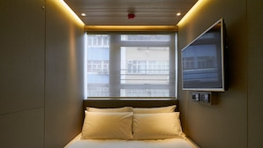 Una habitación de hotel pequeña y moderna con una cama individual, una ventana con persianas, un televisor montado y una lámpara en el techo.