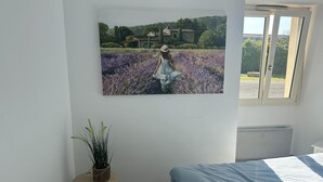 A bedroom with a bed, a window with a view of a house, and a painting of a woman in a field.