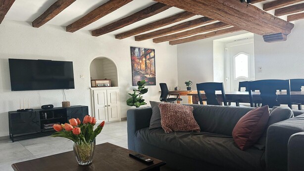 A living room with a flat-screen TV, a sofa, a coffee table with a vase of flowers, and a dining area with chairs and a table.