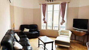 A living room with a black leather sofa, a white armchair, a wooden coffee table, a TV on a wooden stand, and a window with curtains. - Nice center 4-room apartment with air conditioning (Nice)