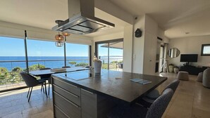 A modern kitchen with a large island, a dining area with chairs, a view of the sea, and a living area with a TV and seating. - Villa Big view (Roquebrune-sur-Argens)