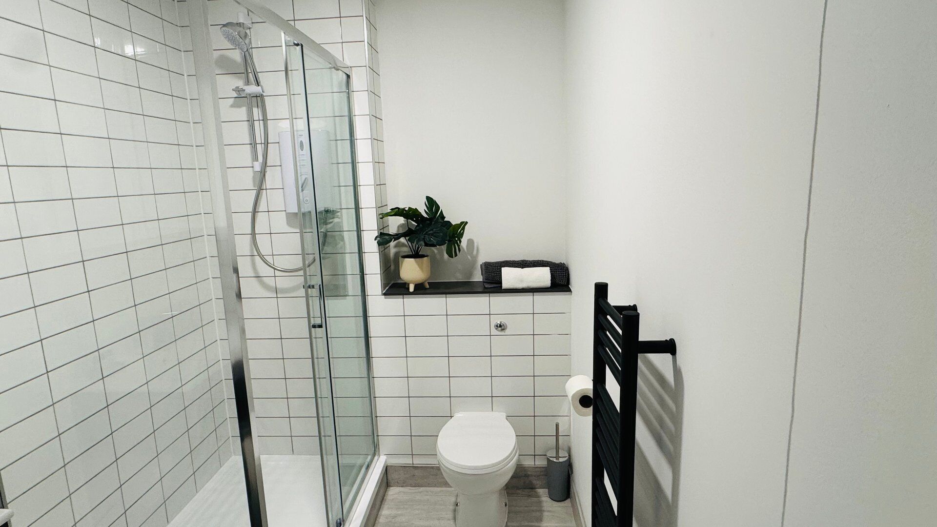 Et badeværelse med hvide fliserede vægge, en glasbrusekabine, et toilet, en vask og en potteplante.
