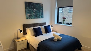 A bedroom with a bed, a nightstand, a lamp, a window with a view, and a painting on the wall. - Well-Located 2-Bed Flat, Swindon Old Town (Swindon)