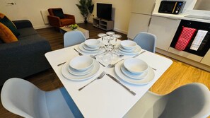 A dining area with a white table set for six, surrounded by blue chairs, a sofa, a TV, and a kitchen with a microwave. - Well-Located 2-Bed Flat, Swindon Old Town (Swindon)