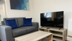 Ein Wohnzimmer mit grauem Sofa, zwei blauen Kissen, einem Holztisch, einem Fernseher auf einem Schrank und einem Bild an der Wand.