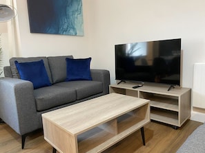 A living room with a grey sofa, two blue cushions, a wooden coffee table, a TV on a stand, and a painting on the wall. - 3-Bed Apartment, Swindon Old Town (Swindon)