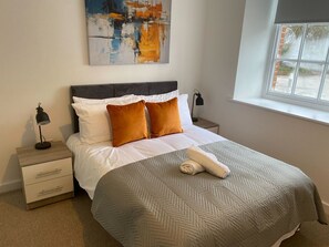 A neatly made bed with a patterned bedspread, two pillows, a bedside lamp, a nightstand, and a window with a view. - 3-Bed Apartment, Swindon Old Town (Swindon)