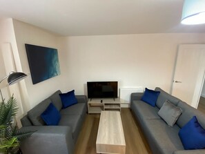 A modern living room with two grey sofas, a coffee table, a flat-screen TV, and a painting on the wall. - 3-Bed Apartment, Swindon Old Town (Swindon)