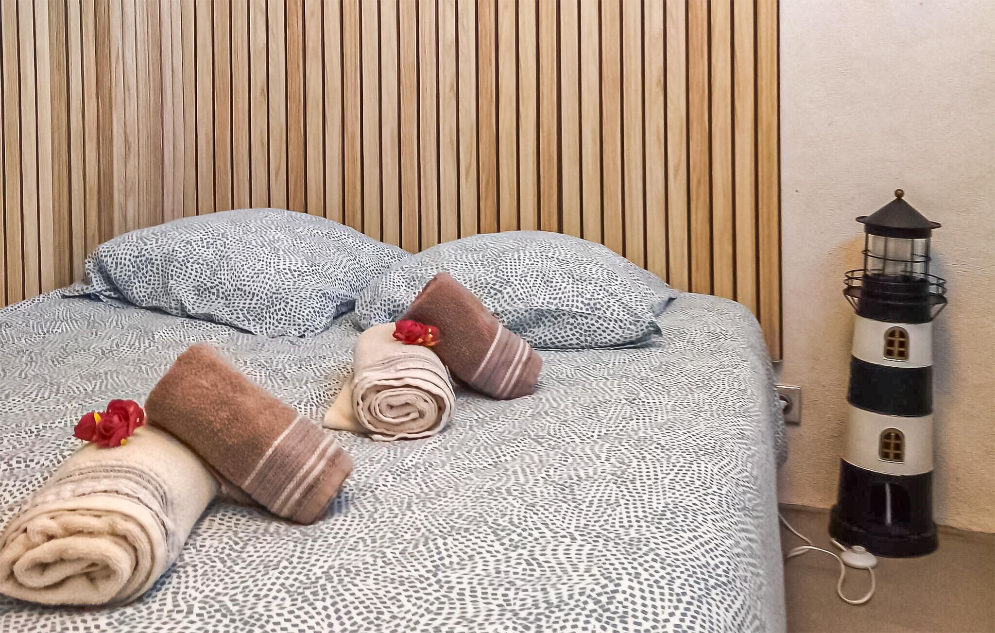 Un lit bien fait, avec une literie à motifs et des serviettes roulées ornées de fleurs, une tête de lit en lattes de bois et une décoration représentant un phare sur le mur.