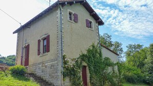 Un bâtiment en pierre de deux étages, avec des volets rouges, un petit escalier à l’avant et un jardin agrémenté de plantes grimpantes.