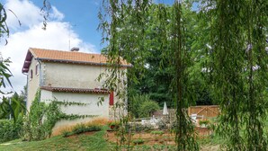 Une maison à deux étages, avec un toit recouvert de tuiles, entourée d’un jardin où poussent différentes plantes et fleurs.