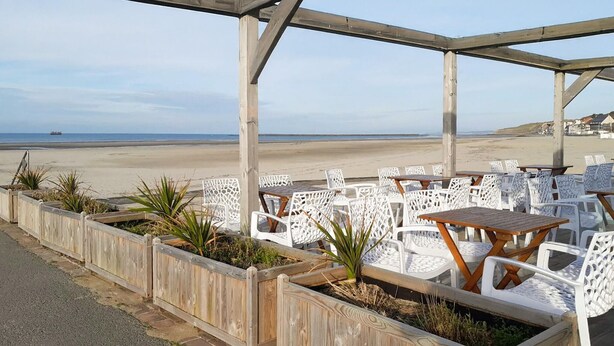Eine Terrasse direkt am Strand mit weißen Korbstühlen und Holztischen, mit Blick auf den Strand und den Ozean.