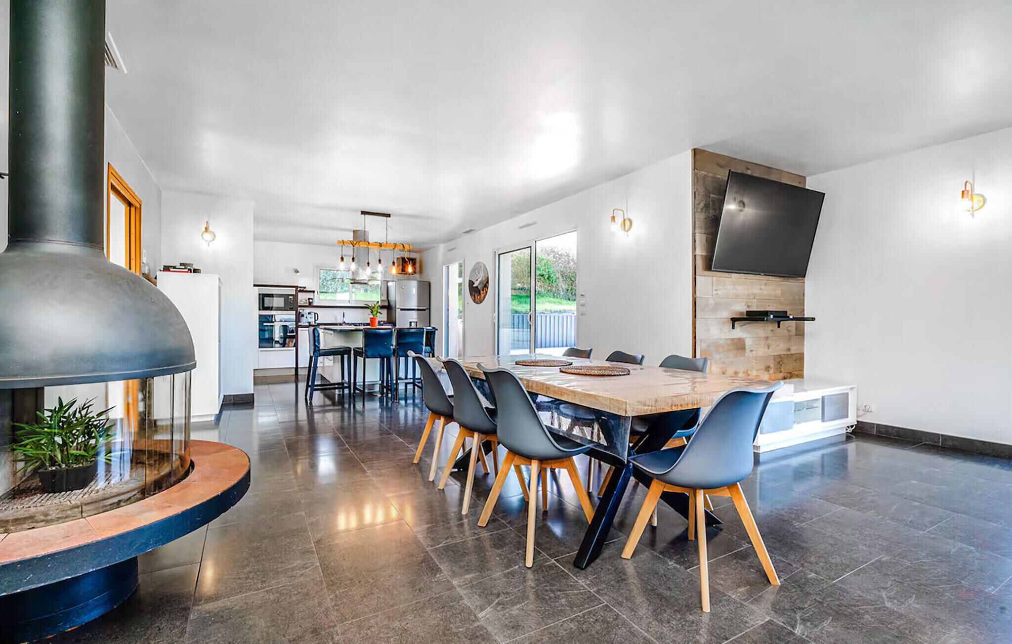 A modern kitchen with a large island, a dining area with a long table and chairs, and a TV mounted on the wall.