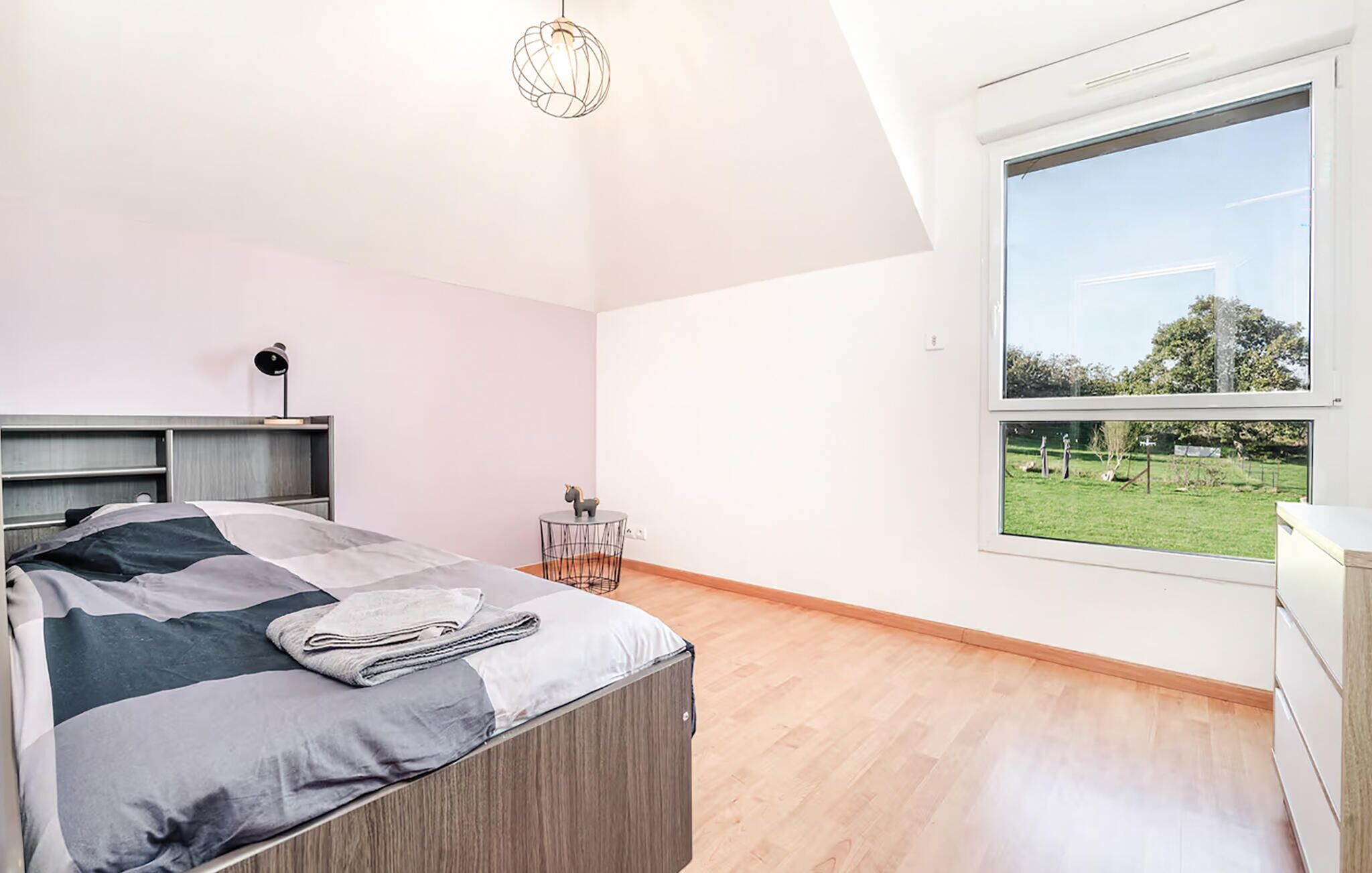 A modern bedroom with a large window, a bed with a grey and white duvet, a small round table with a chair, and a desk with a lamp.