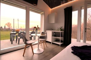 A modern room with a large window, a dining area with chairs and a table, a bed with a purple blanket, and a small white cabinet. - Nature Escape Villa Pool (Montebelluna)