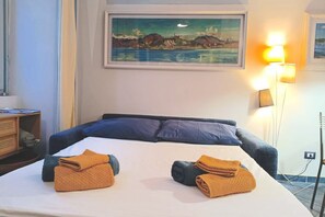 A hotel room with a bed, pillows, a painting on the wall, a lamp, and a desk with a chair. - Charming one-bedroom apartment, finely furnished, located on the ground floor of Via Pertossi in the historic centre of Arona.Casa Azzurra is a 52 sqm one-bedroom apartment with air conditioning, featuring a living room/kitchen area with a double sofa be (Arona)
