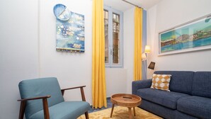 A room with a blue sofa, a blue armchair, a round wooden coffee table, yellow curtains, a window with a view, and a painting on the wall. - Charming one-bedroom apartment, finely furnished, located on the ground floor of Via Pertossi in the historic centre of Arona.Casa Azzurra is a 52 sqm one-bedroom apartment with air conditioning, featuring a living room/kitchen area with a double sofa be (Arona)