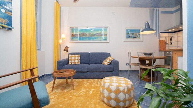 A living room with a sofa, a round ottoman, a coffee table, and a dining area with a table and chairs. - Charming one-bedroom apartment, finely furnished, located on the ground floor of Via Pertossi in the historic centre of Arona.Casa Azzurra is a 52 sqm one-bedroom apartment with air conditioning, featuring a living room/kitchen area with a double sofa be (Arona)
