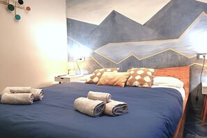 A hotel room with a bed, pillows, a nightstand, a lamp, and a wall with a geometric design.