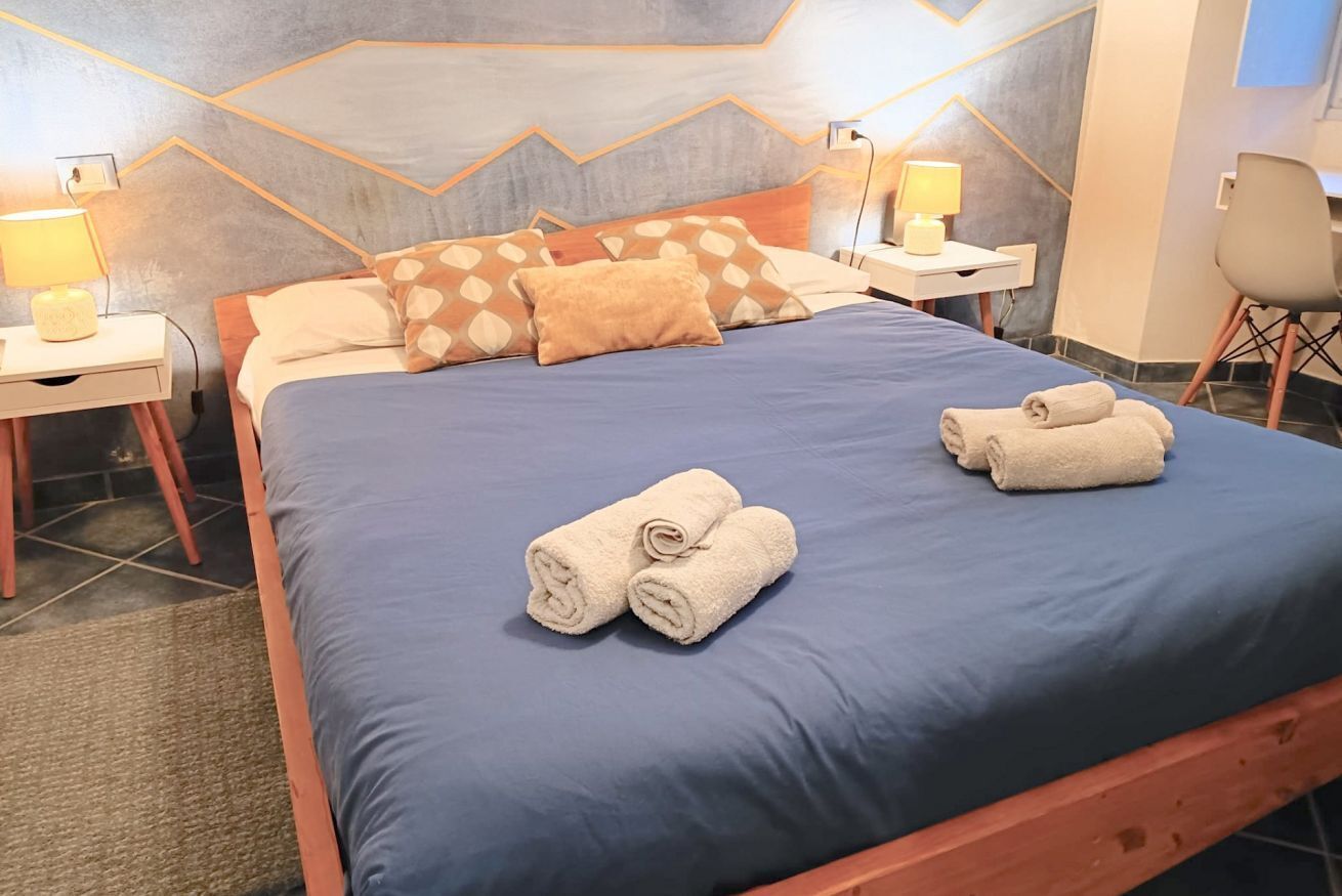 Ein ordentlich bezogenes Bett mit blauer Bettwäsche, zwei Nachttische mit Lampen und ein Sessel in einem modernen Zimmer mit geometrischem Wanddesign.