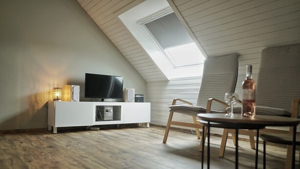Un salon moderne doté d’un plafond en pente, d’un téléviseur, d’un meuble de rangement blanc pour appareils multimédias, d’un plancher en bois et d’un coin repas avec une table et des chaises.