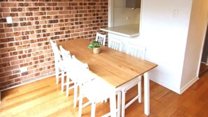 Un coin repas avec une table en bois et des chaises blanches, un mur en briques et une fenêtre.
