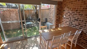 Une pièce comprenant une table à manger en bois, des chaises blanches, une porte-fenêtre coulissante donnant sur une cour arrière avec un mur en briques, et un luminaire suspendu.