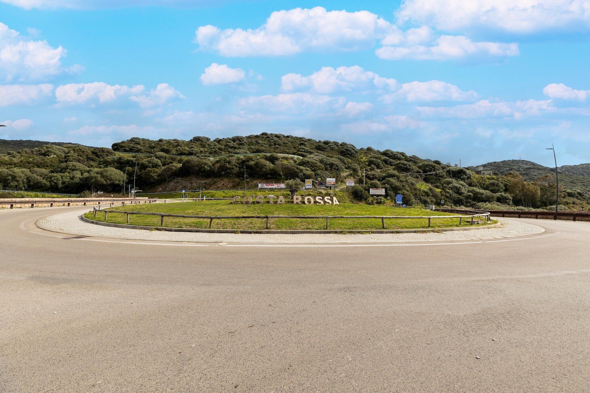 A roundabout with a sign reading 'POSTA ROSSA' and a grassy area with a fence.