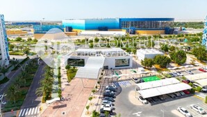 A modern building complex with multiple structures, including a large blue building and a white building with a curved roof. There are parking areas and a road with palm trees. - Bloomfields 1br Deluxe in Water Edge (Abu Dhabi)