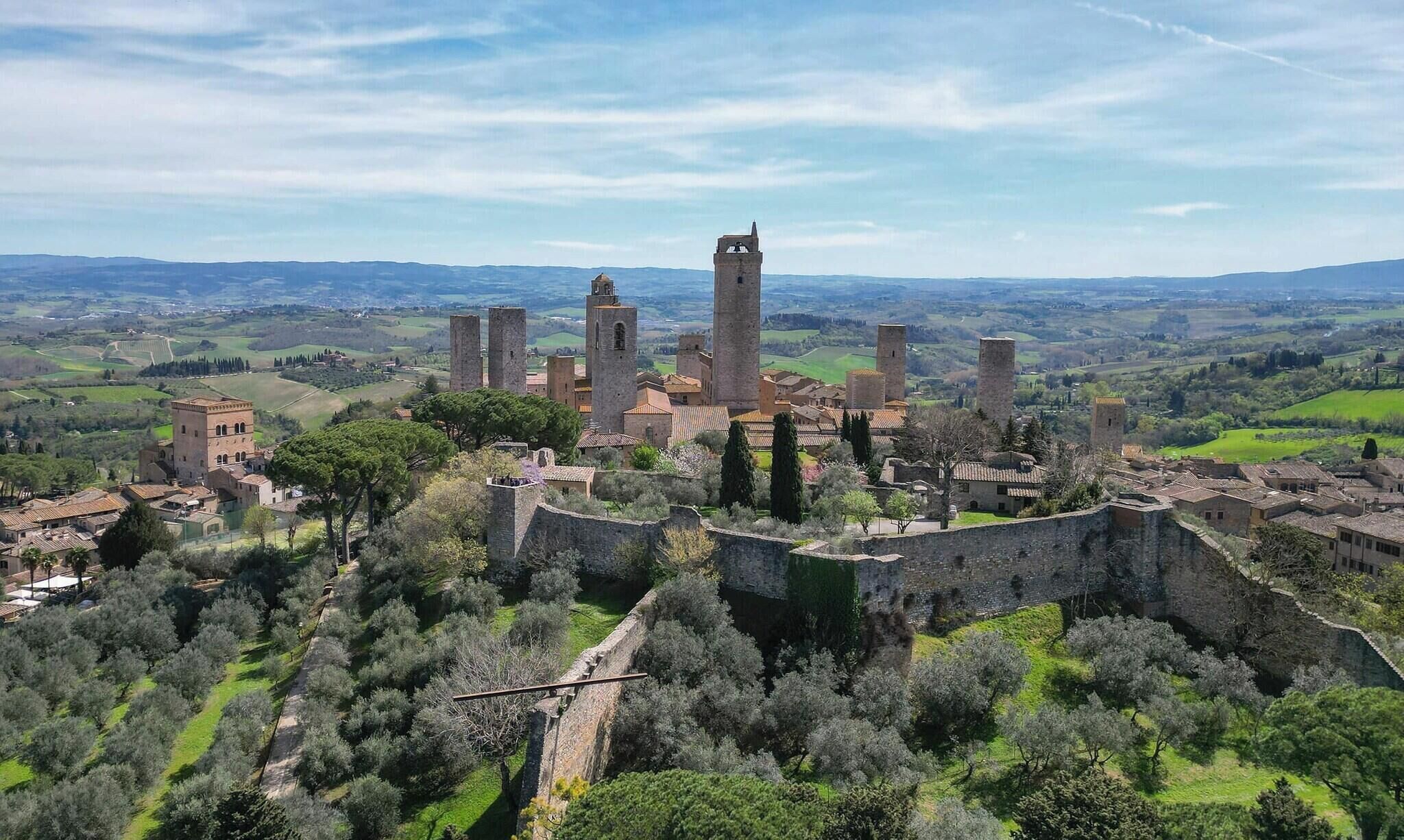 A historic fortified town with prominent towers and a central bell tower, surrounded by olive groves and rolling hills.