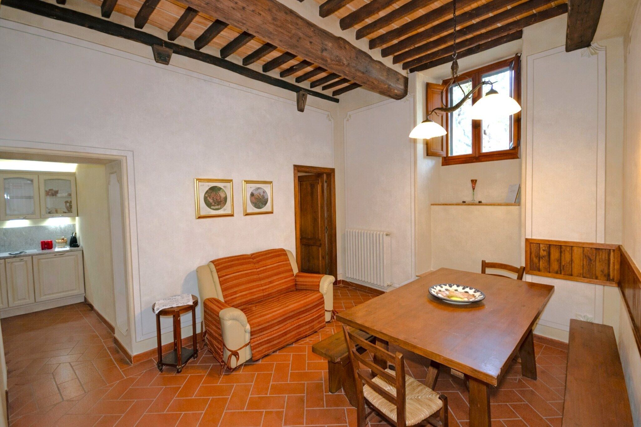 A cozy dining area with a wooden table, chairs, and a bench. There is a sofa and a small table with a lamp. The room has a rustic ceiling with exposed beams and a window with a wooden frame.