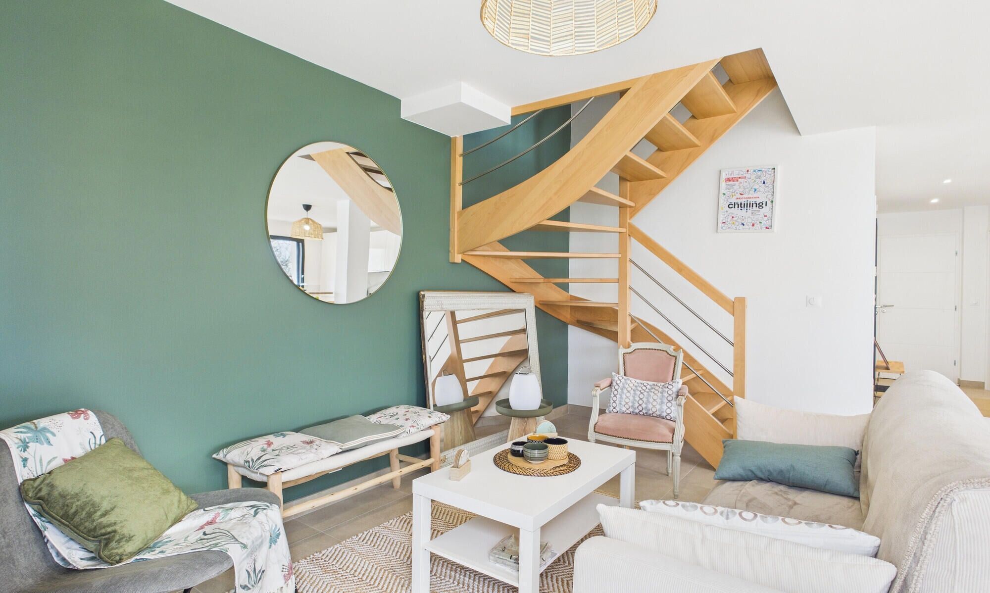 Ein modernes Wohnzimmer mit einer Holztreppe, einem runden Spiegel, einem Sofa, einem Sessel und einem Couchtisch.
