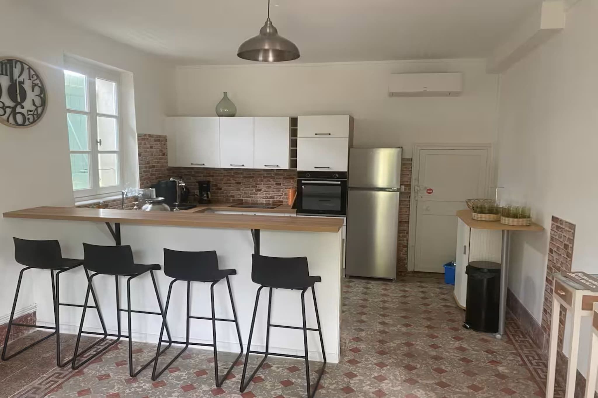 Una cocina con una isla central, taburetes de bar, un reloj y un refrigerador.