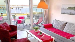 A cozy living area with a sofa, red armchair, and a table with a red table runner. A large window offers a view of a pier and water. - Coastal Apartment with Balcony (Fehmarn OT Burgtiefe-Südstrand)