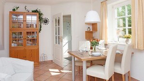 Un comedor con una mesa de madera, sillas blancas y una lámpara colgante blanca. Hay un armario con puertas de vidrio y una puerta que da a otra habitación.