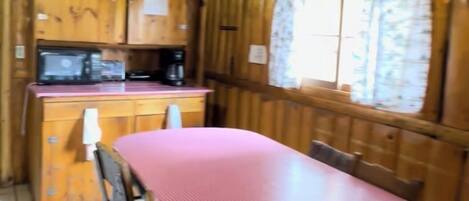 L’intérieur d’un chalet en bois comprend une table à manger, des chaises, un coin cuisine et une fenêtre avec des rideaux.