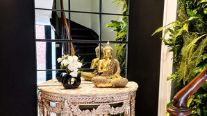 Eine Treppe mit Geländer, ein runder Tisch mit Buddha-Statue und Blumenvase, und ein großer Spiegel, der die Treppe und eine Ziegelwand reflektiert.
