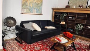 A living room with a black sofa, a red patterned rug, a wooden coffee table with books and a toy train, a map on the wall, and a bookshelf with various items. - Chic vintage apartment in Spa (Spa)