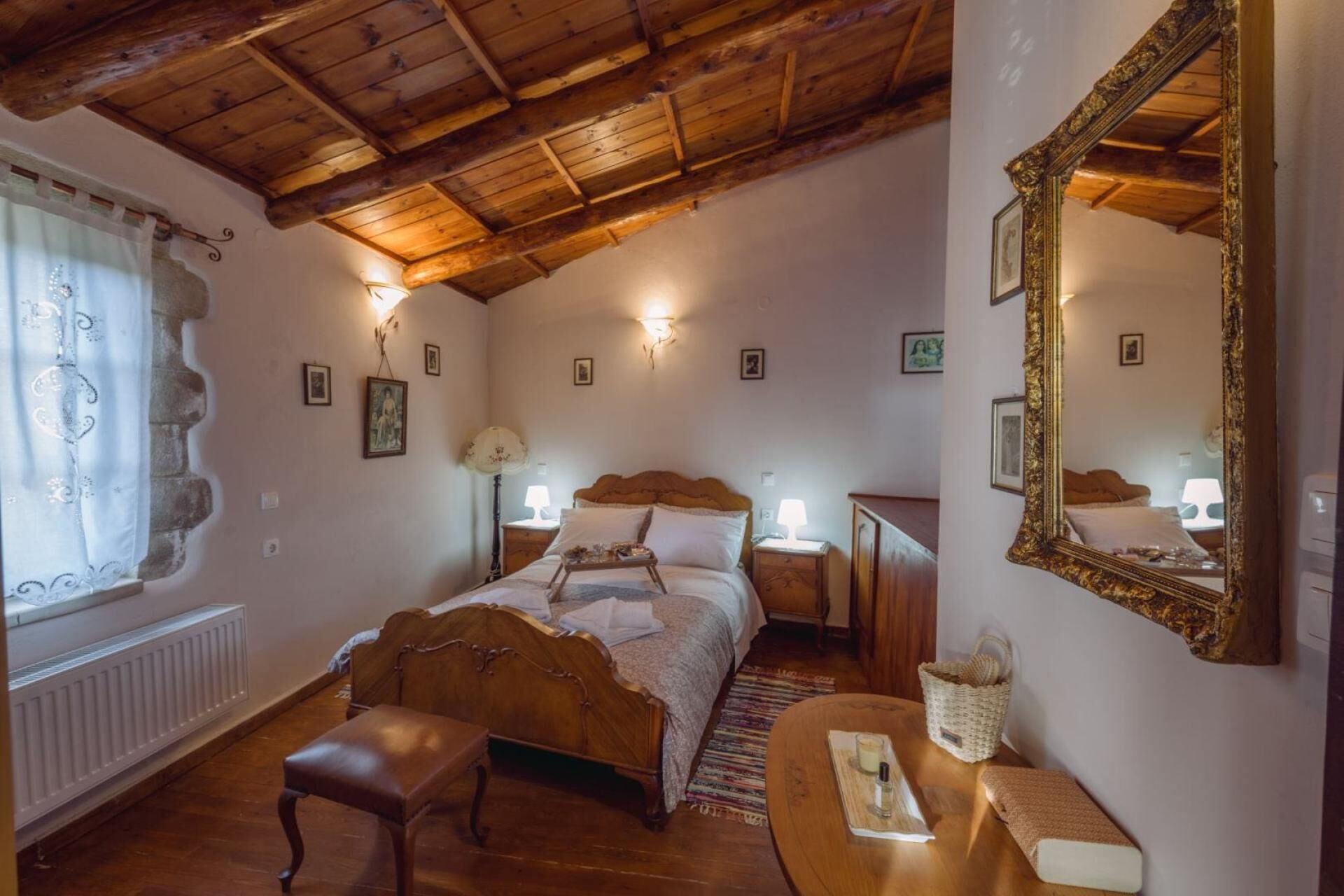 A cozy bedroom with a wooden ceiling, a large ornate mirror, a bed with a wooden headboard, bedside tables with lamps, and a window with curtains.