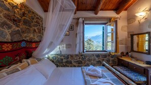 Un dormitorio con pared de piedra, cama con ropa blanca, ventana con vista a las montañas y techo de madera.