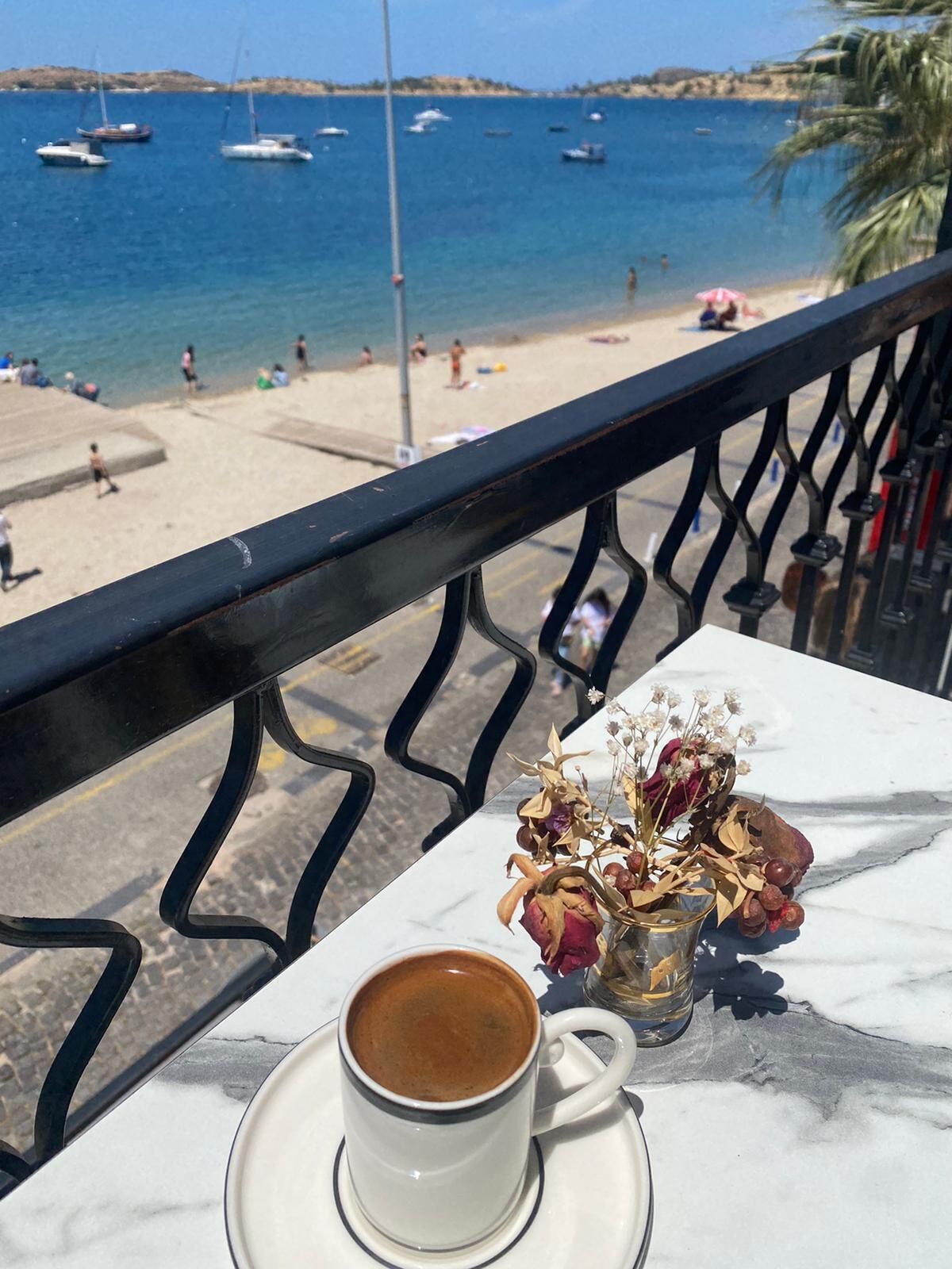 A table with a cup of coffee and dried flowers, overlooking a beach with boats and people.