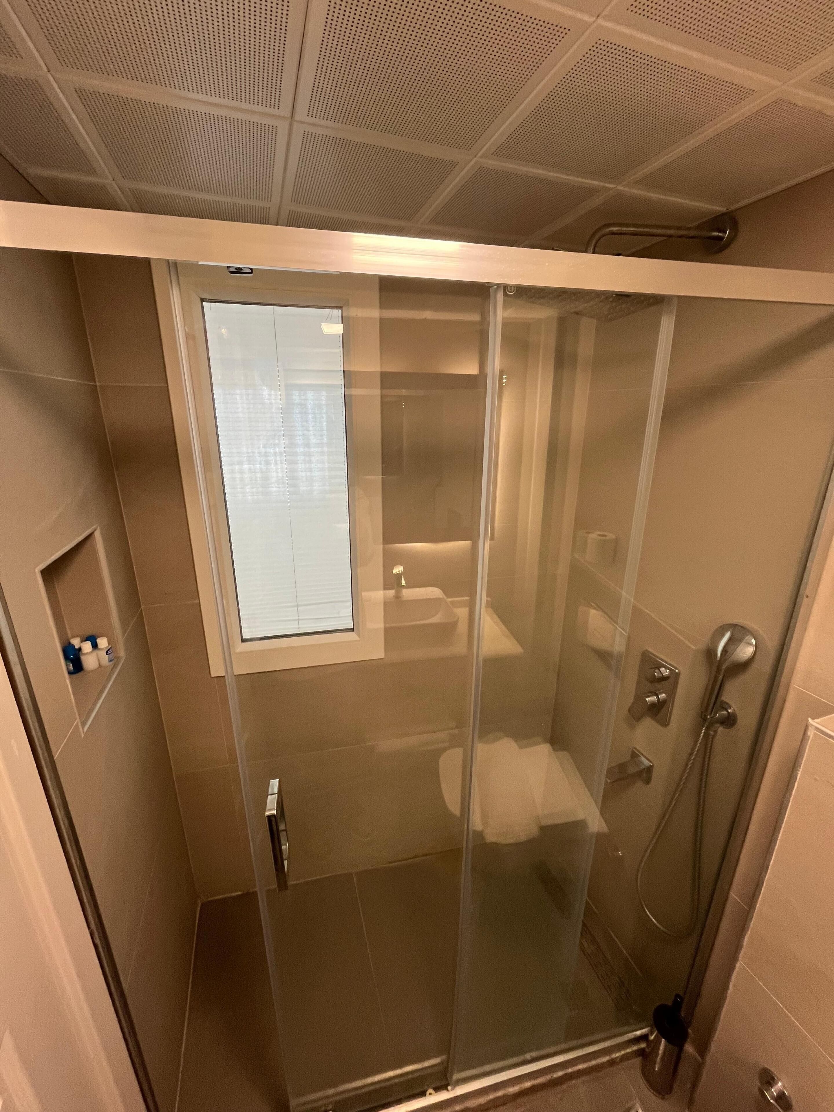 Une cabine de douche fermée par des parois en verre, avec des toilettes et un lavabo, une petite étagère pour les articles de toilette et une bouche d’aération au plafond.