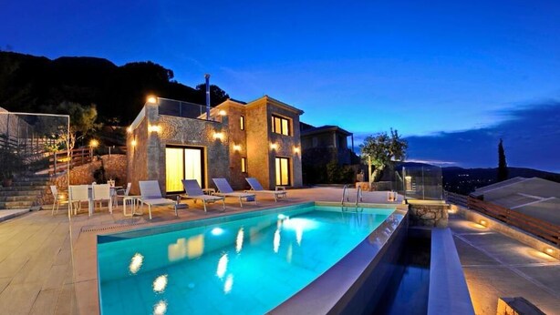 A modern pool area with a rectangular pool, lounge chairs, and a stone building with warm lighting. - Stone Crafted Luxury Villas - Villa Artemis (Nikiana)