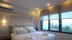 A modern bedroom with a large bed, a headboard with a patterned design, bedside lamps, and a view through a window. - Stone Crafted Luxury Villas - Villa Artemis (Nikiana)