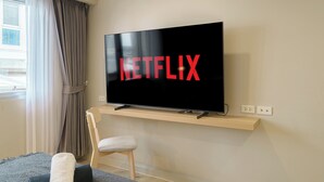 A modern living room with a mounted TV displaying the Netflix logo, a wooden shelf below, a window with curtains, and a chair. - Casa Amor (Bangkok)